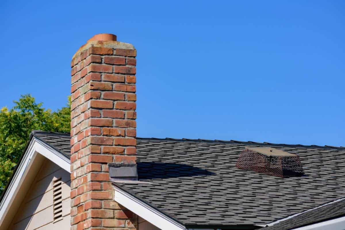 A close-up of a house rooftop with dark shingles, featuring a tall red brick chimney and a metal-caged vent against a clear blue sky. Green tree foliage is partially visible in the background.