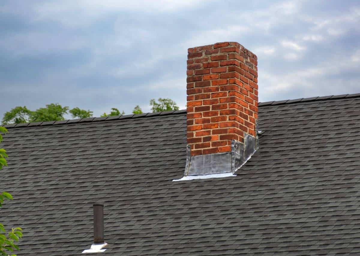 A brick chimney extends from the gray shingled roof of a house, surrounded by trees and under a cloudy sky.