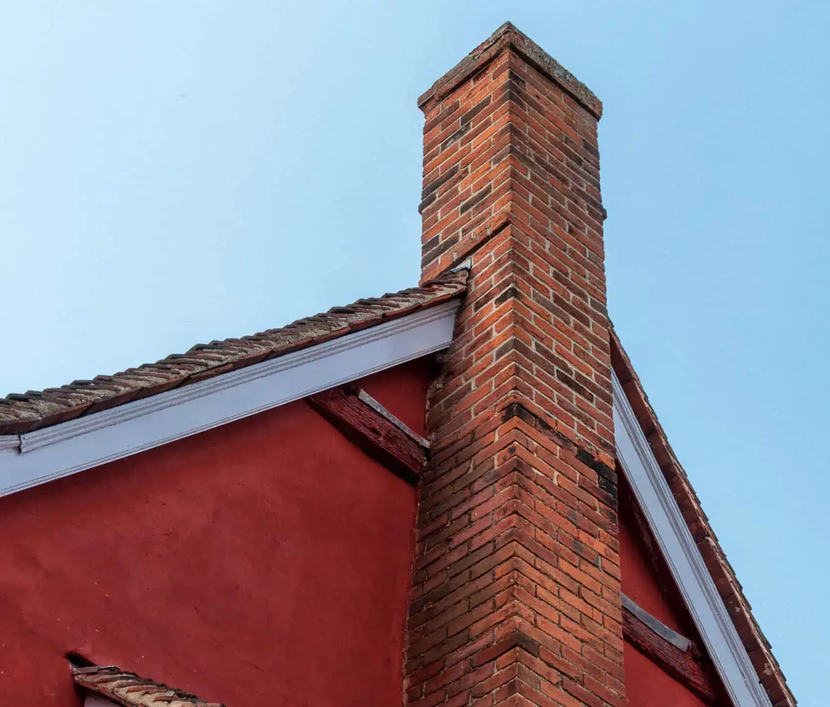 A red-brick chimney extending from the tiled roof of a red-walled building under a clear blue sky, showcasing architectural details and construction materials.